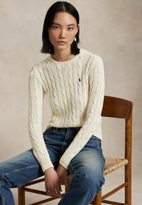 Femme aux cheveux noirs et raides portant un pull torsadé crème et un jean bleu, assise sur une chaise en bois sur un fond neutre.