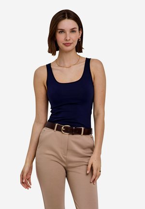 Mujer con cabello corto y castaño, vestida con una camiseta sin mangas azul marino, pantalones beige y un cinturón marrón oscuro, de pie contra un fondo liso y claro.