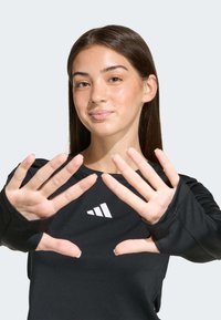 Čierne športové tričko s dlhým rukávom má na hrudi biele logo, otvory na palce na rukávoch a hladkú, pružnú textúru látky.