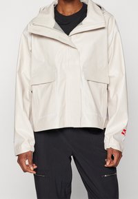 Veste imperméable beige clair avec capuche, deux poches avant à rabat, poignets élastiques et accents réfléchissants. Texture lisse et coupe courte.