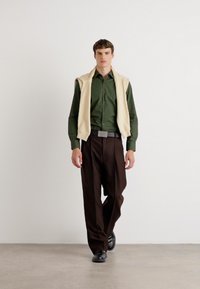 Modèle portant une chemise boutonnée vert foncé, un pull beige drapé sur les épaules, un pantalon plissé marron et des chaussures noires sur un fond uni.