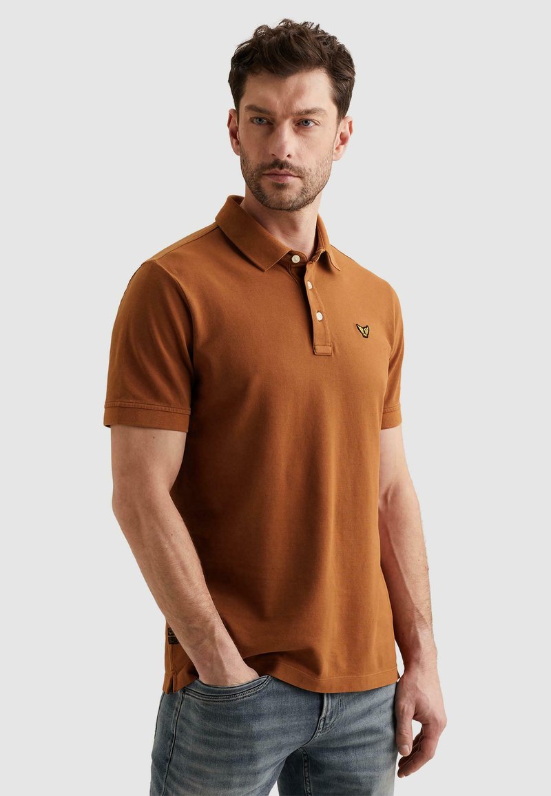 Homme portant un polo à manches courtes marron avec des boutons et un logo, associé à un jean bleu, debout avec une main dans la poche.