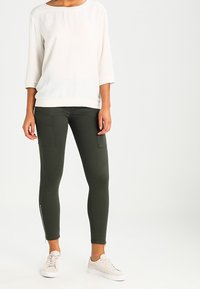 Blusa blanca de corte holgado con mangas de tres cuartos, combinada con pantalones ajustados de color verde oscuro que cuentan con bolsillos laterales y cremalleras en los tobillos.