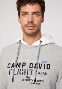 Felpa grigia con interno bianco, caratterizzata da un testo ricamato in nero e blu. Ha un cordino con una texture intrecciata e punte in metallo.