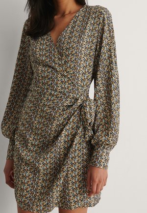 Mujer vestida con un mini vestido de manga larga y estilo cruzado con un pequeño estampado floral en tonos amarillos, azules, blancos y negros suaves.