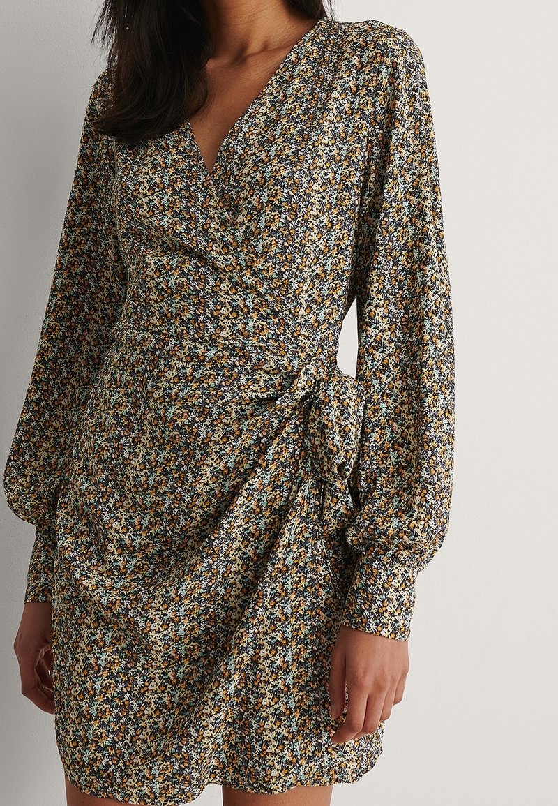 Femme portant une mini-robe portefeuille à manches longues avec un petit imprimé floral dans des tons jaunes, bleus, blancs et noirs atténués.