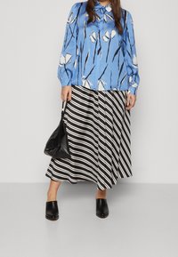 Donna che indossa una blusa floreale blu, una gonna midi a righe bianco e nere, mule nere e tiene una borsa nera, in piedi su uno sfondo neutro.