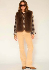 Gilet en fausse fourrure marron sur une chemise à carreaux, associé à un pantalon évasé en velours côtelé beige et des bottes à talons marron. Accessoirisé avec des lunettes de soleil.