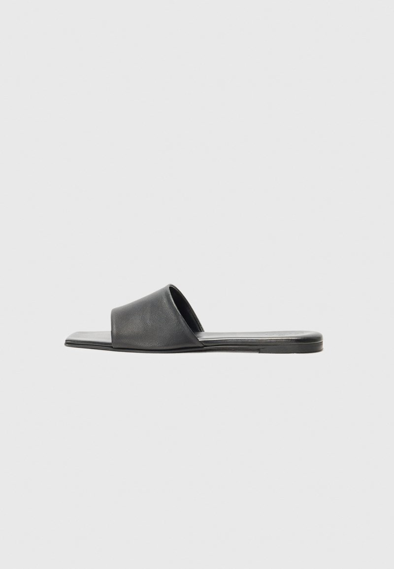 Sandale à glisser en cuir noir avec une large sangle unique et une semelle plate, présentant une texture lisse et un design minimaliste.