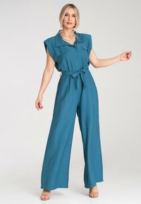 Szmaragdowy kombinezon z szerokimi nogawkami, krótkimi rękawami, guzikiem z przodu i paskiem w talii. Lekki materiał o gładkiej, matowej fakturze.