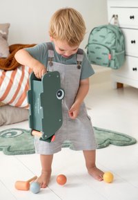 Groene speelopbergdoos met ogen, oranje handvatten; kind in grijze tuinbroek houdt het vast. Kleurrijke ballen liggen op de vloer, en een pluche vloerkleed is zichtbaar.