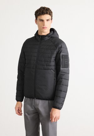 Jonge man in een zwart gewatteerd jack met capuchon, voorzien van ritszakken en een logopatch op de bovenste linkerarm, gecombineerd met grijze broek.