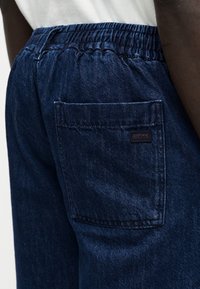 Σκούρο μπλε τζιν παντελόνι με ελαστική μέση και πίσω τσέπη με μικρή ετικέτα "BOSS" σε άτομο που φοράει λευκό τοπ.