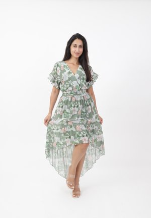 Robe fluide verte avec motif floral, décolleté en V et manches à volants. Ourlet asymétrique et tissu transparent. Associée à des sandales à talons transparents.