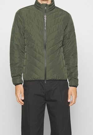 Mężczyzna w zielonej pikowanej kurtce z wysokim kołnierzem i czarnym zamkiem błyskawicznym, w czarnych spodniach, stojący na jednolitym tle.