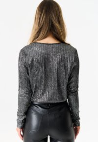 Woman with long hair wearing a shiny silver ribbed long-sleeve top and black leather pants, shown from the back against a white background.