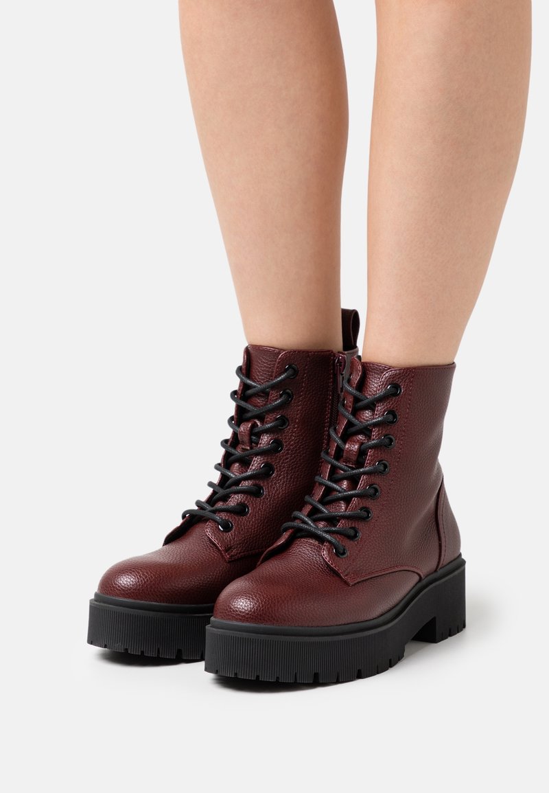 Bottes en cuir bordeaux montant à la cheville avec semelles épaisses à crampons noires et lacets noirs, portées sur des jambes nues, sur fond blanc.