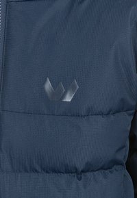Marinblå puffjacka med texturerad tyg, med en metallisk logotyp. Designen inkluderar quiltade sektioner och en sömlös dragkedja.