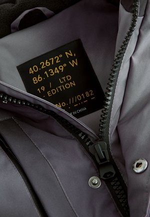 Vnútro sivej syntetickej bundy s čiernym zipsom. Textilná štítok zobrazuje súradnice, "19/LTD EDITION" a "No. ///0182".