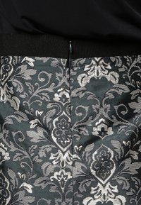 Tissu brocart noir et argent avec un motif damassé floral complexe, gros plan d'une couture zippée sous une taille noire.