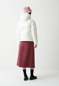 Vit puffjacka med huva, rosa bucket hat, vinröd kjol, svarta strumpor och svarta skor, sedd bakifrån mot en neutral bakgrund.