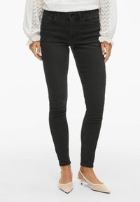 Zwarte skinny jeans met een gladde textuur en een middenris taille, gecombineerd met lichtroze puntige hakken, die een eigentijdse pasvorm uitstralen.