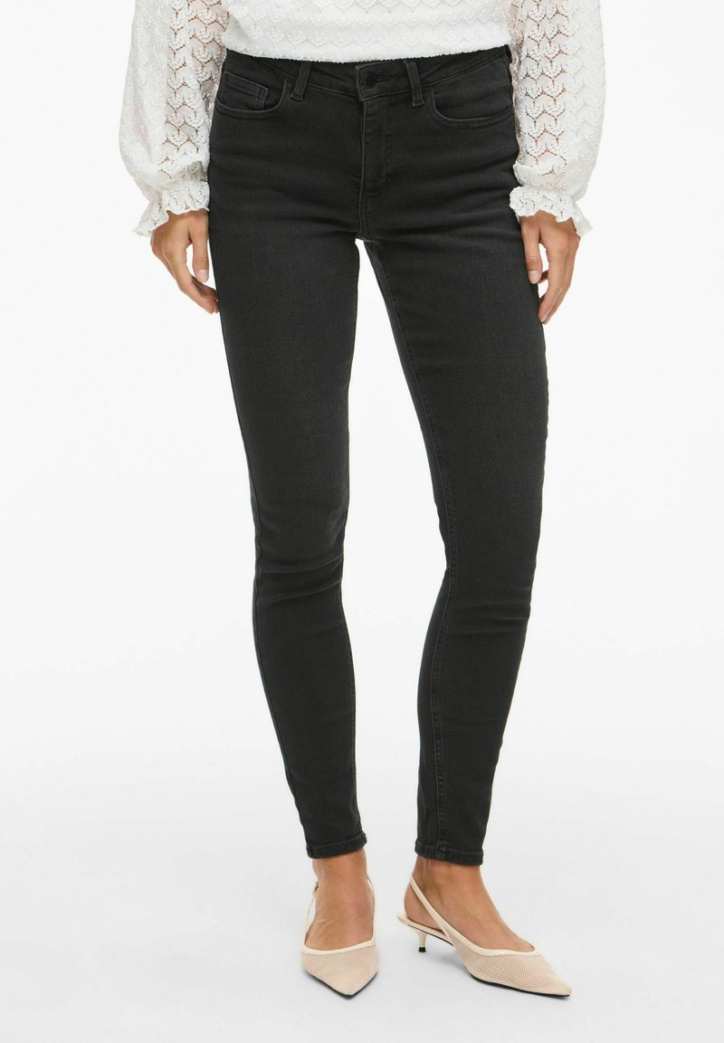 Zwarte skinny jeans met een gladde textuur en een middenris taille, gecombineerd met lichtroze puntige hakken, die een eigentijdse pasvorm uitstralen.