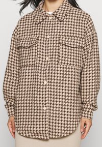 Hnědo-beige kostkovaná košile s límečkem, dvěma náprsními kapsami a zapínáním na knoflíky. Měkká, texturovaná tkanina, dlouhé rukávy.