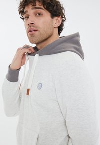 Felpa con cappuccio grigio chiaro con cappuccio grigio scuro, dotata di una tasca frontale, cordini bianchi e un logo rotondo sul petto. Tessuto morbido.