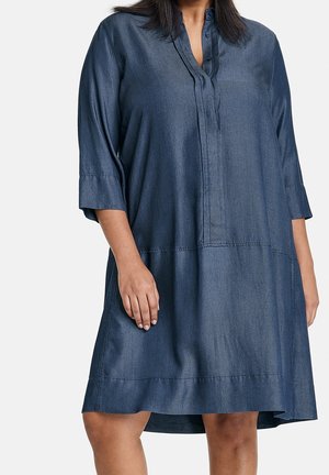 Nainen, joka pukeutunut väljästi istuvaan farkkumekkoon, jossa on kolme neljäsosa hihaa ja V-pääntie, seisomassa valkoista taustaa vasten.