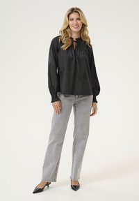 Blusa nera a maniche lunghe con motivo floreale, scollatura arricciata e dettaglio con fiocco, abbinata a jeans dritti grigi chiari e tacchi neri.