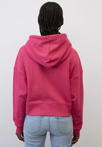 Persona con trenzas, vistiendo un sudadera rosa y jeans de un azul claro, de pie frente a una pared blanca lisa.