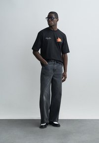 Mees mustade prillide, musta graafilise t-särgi, laia säärega tuhmide mustade teksade ja must-valgete kingadega, seisab ühe käega taskus.