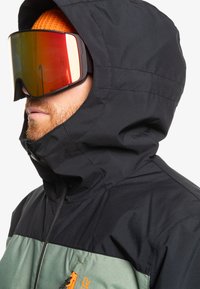 Chaqueta negra con capucha y panel inferior verde, detalle del zipper en naranja, y una persona usando gafas de esquí de gran tamaño con espejo. Textura suave.