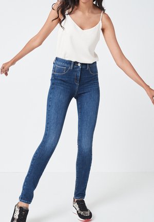 Skinny jeans in dark blue denim with five pockets, light stitching, and high waist. Worn with a white spaghetti strap top and patterned sneakers.
