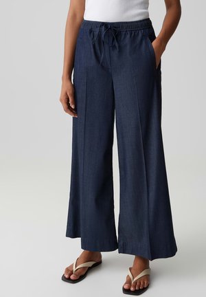 Frau trägt weite marineblaue Hosen mit Kordelzugbund, weißes Tanktop und beige Flip-Flops, steht mit einer Hand in der Tasche.