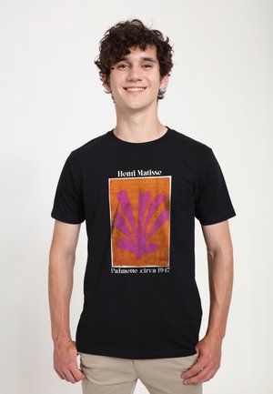 Ung mand med krøllet hår smiler iført en sort T-shirt med Henri Matisse Palmette-kunstværk i lilla på orange baggrund med beige bukser.