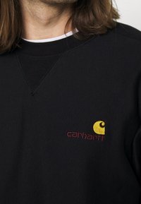 Schwarzer Sweatshirt mit Rundhalsausschnitt, das ein gelb-rotes gesticktes Logo auf der Brust trägt. Das Material wirkt glatt mit einer leichten Textur.