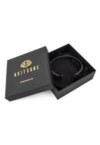 Boîte à bracelet noire ouverte avec logo Akitsune doré, contenant un bracelet manchette noir avec des têtes de renard aux extrémités, sur une doublure en tissu noir.
