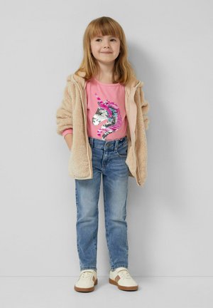 Jong meisje draagt een beige fleecejack met ritssluiting, een roze shirt met een pailletten eenhoorn, een blauwe spijkerbroek en witte sneakers, staand tegen een grijze muur.