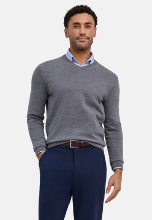 Homme portant un pull gris à col en V par-dessus une chemise bleu clair à col, avec un pantalon bleu marine et une ceinture marron, debout avec une main dans la poche.