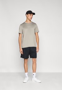 Ανοιχτό γκρι κοντομάνικο μπλουζάκι με λογότυπο, συνδυασμένο με σκούρα σορτς από διχτυωτό ύφασμα. Λευκά αθλητικά παπούτσια και μαύρο καπέλο ολοκληρώνουν το αθλητικό look.