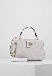 Petit sac à main beige et blanc Guess avec motif monogramme, plaque logo dorée, anse supérieure et bandoulière amovible sur surface blanche.