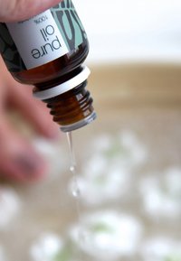 Klar glassflaske med hvit kork merket "Pure Oil," som heller en klar væske. Bakgrunnen viser uskarpe hvite og grønne elementer.
