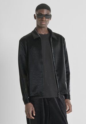 Zwarte jas met krokodillenpatroon, ritssluiting aan de voorkant en een puntige kraag, gecombineerd met een zwart t-shirt met kleine versieringen.