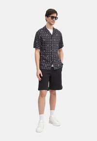 Camisa de manga corta, tipo botonera, en negro con patrón geométrico blanco, combinada con pantalones cortos negros y zapatillas blancas. Modelo con gafas de sol.