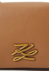 Borsa in pelle sintetica marrone con un accento dorato del logo, caratterizzata da linee fluide e stilizzate che creano un moderno dettaglio visivo sulla superficie.