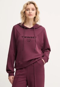 Bordová mikina s kapucňou a dlhými rukávmi, s kontrastným prešívaním a predným logom. Mäkko tkanina a uvoľnený strih.