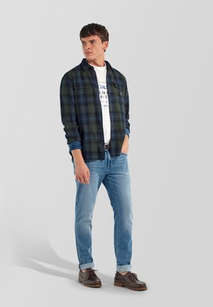 Joven con una camisa de cuadros oscura sobre una camiseta blanca con diseño gráfico, jeans azul claro y zapatos marrones con cordones, de pie frente a un fondo liso.