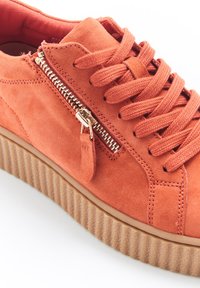 Baskets en suède orange avec une semelle plateforme texturée et un détail de fermeture éclair doré sur le côté, accompagnées de lacets plats à l'avant.